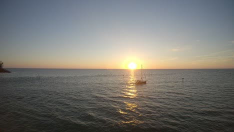 a boat crossing the sea in front of the sun setting over the horizon. drone footage moves backwards captur8ng this beautiful scene and shows some of the beach and surrounding trees.