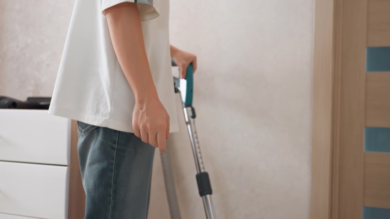 Person vacuuming home hallway floor, canister vacuum blue body, moving along baseboard and dresser, casual jeans white shirt, natural light, quiet morning cleaning routine, focused household chore