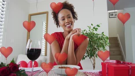 animation of heart balloons over african american woman having video call and drinking wine