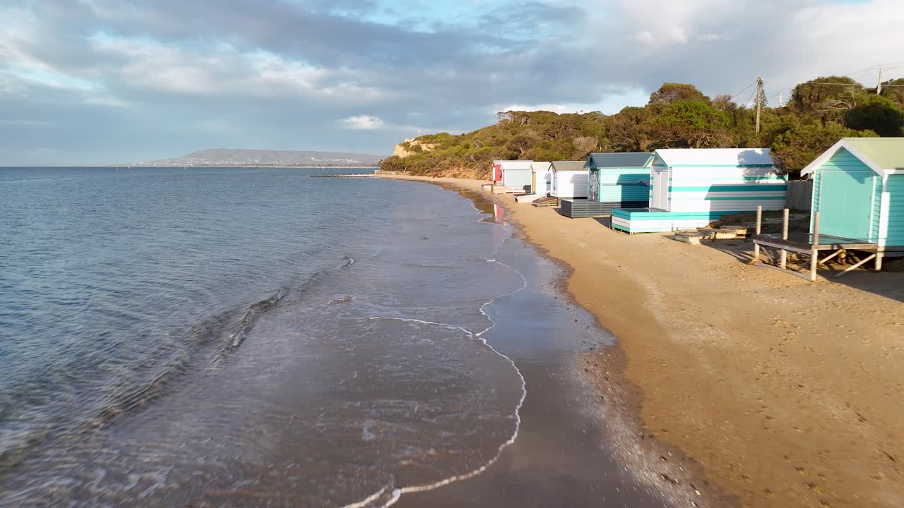 Drone glides above sandy beach, passing vibrant bathing boxes under soft morning sunlight, calm atmosphere