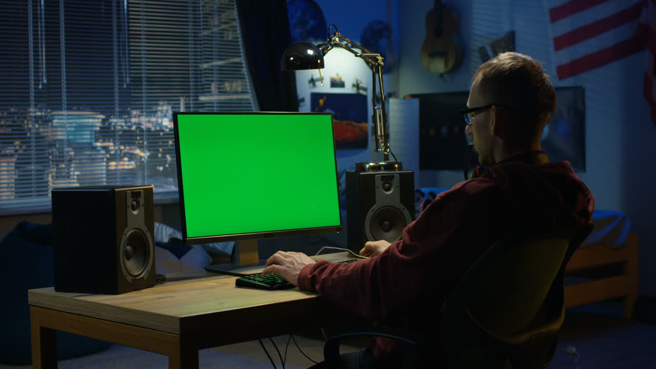 Man working on a computer with a green screen in his bedroom at night