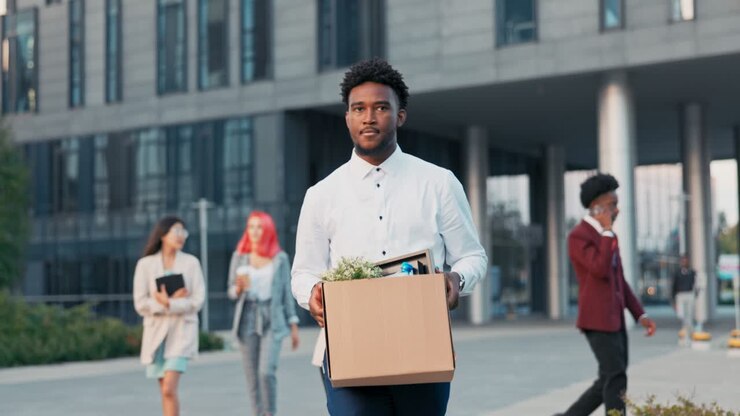 A dark-skinned man long-time office worker is fired from job, walks out of company building with co-workers, holding box of packed belongings in hands, a disgruntled expression, anger, sadness