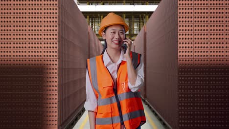 asian female engineer with safety helmet talking on smartphone while standing with red brick packed in stacks are stored