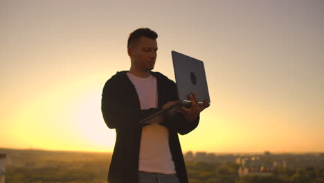 A-man-with-a-laptop-at-sunset-stands-on-the-roof-of-the-building-and-writes-a-code-on-the-keyboard.-Hacker-with-laptop.