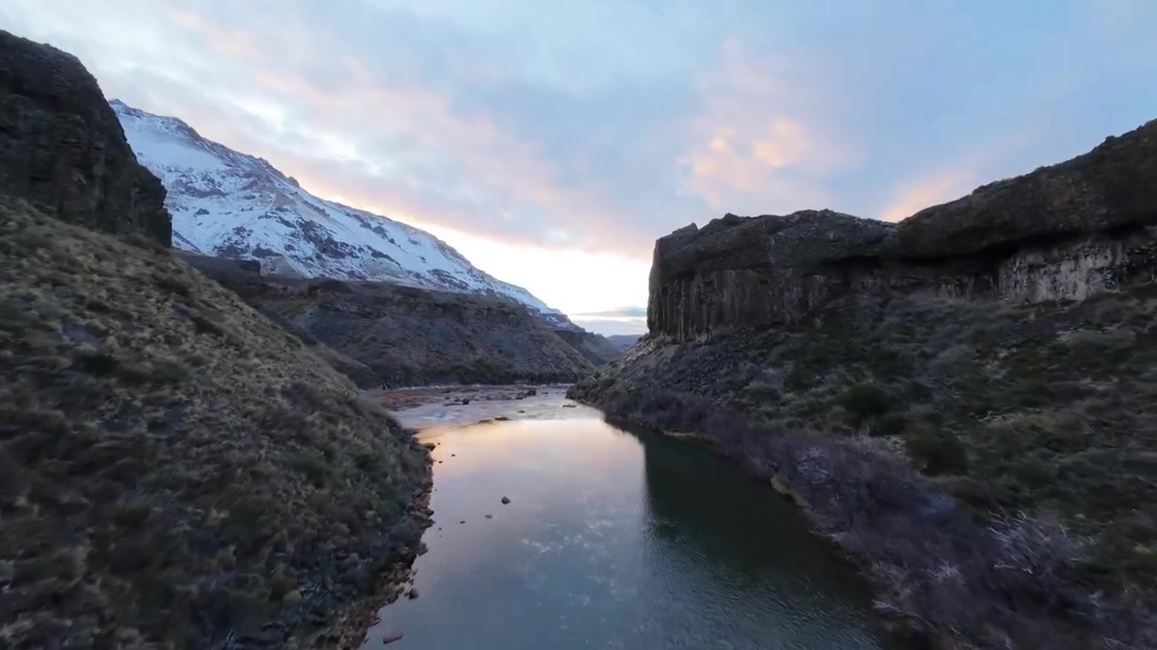 Immersione FPV sopra scogliere rocciose di basalto e le correnti impetuose del fiume che scorrono alla cascata Salto del Agrio all'alba, con un paesaggio invernale innevato circostante.