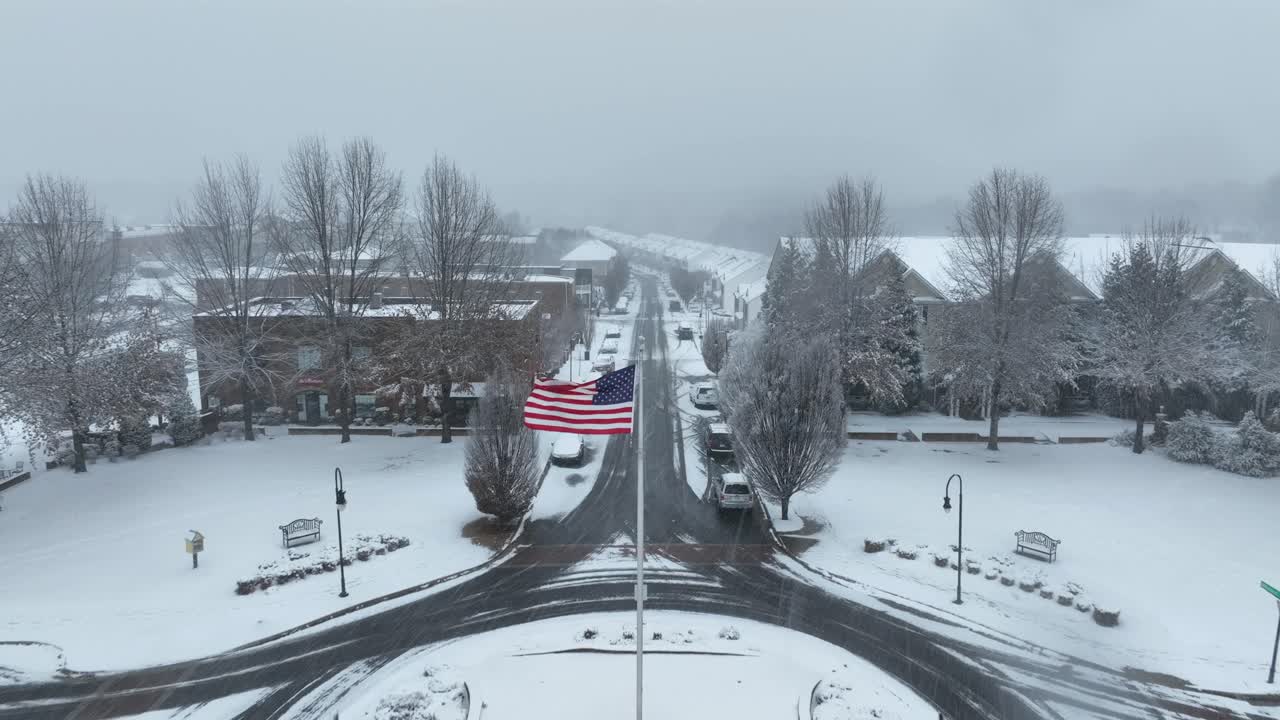Aerial approaching shot of blowing American flag on flagpole of roundabout. Snowscape and snowfall in American town. Dolly forward shot. Housing area of neighborhood in winter