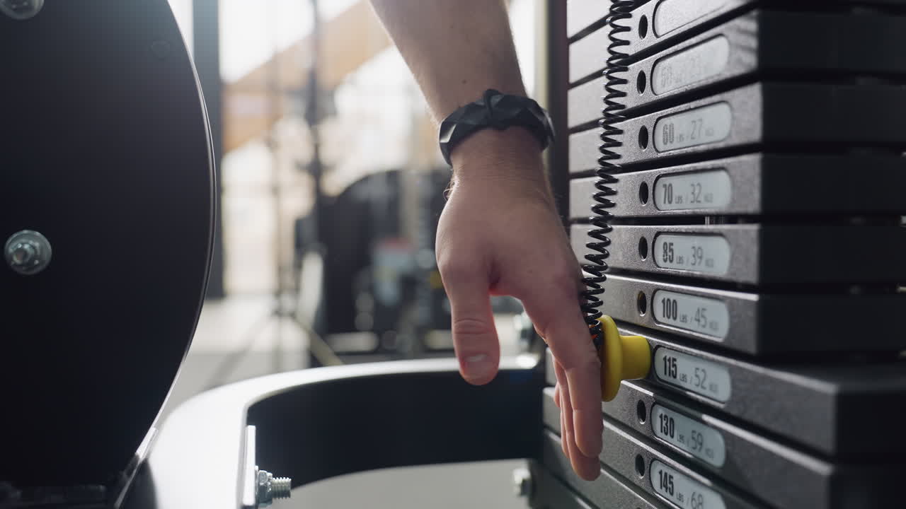 Trainer selects weight by inserting pin into lower plate of stack on cable machine, showcasing precise adjustment process in modern gym setting before workout begins to ensure correct resistance