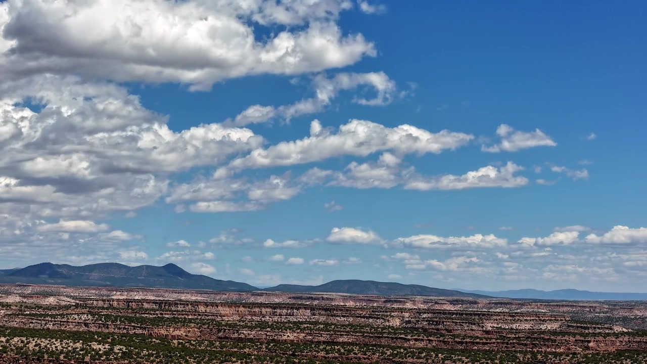 Timelapse 4K UHD con drone di nuvole bianche che si muovono rapidamente attraverso un cielo blu vivido. Ombre dinamiche delle nuvole si muovono rapidamente attraverso i distinti strati orizzontali di un aspro paesaggio di mesa desertica d'alta quota del Nuovo Messico.
