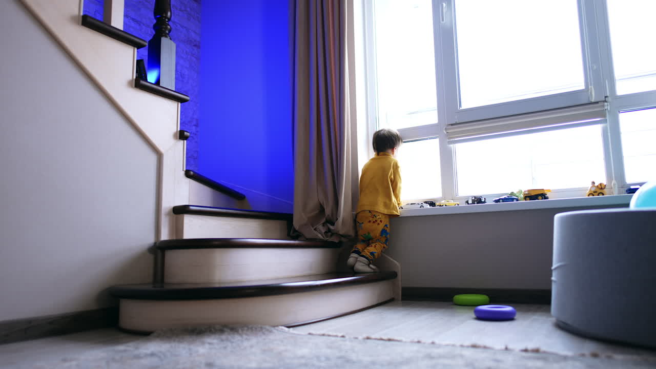 Rear view of a baby boy in yellow clothes standing near the window. Kid stands still looking at the street.