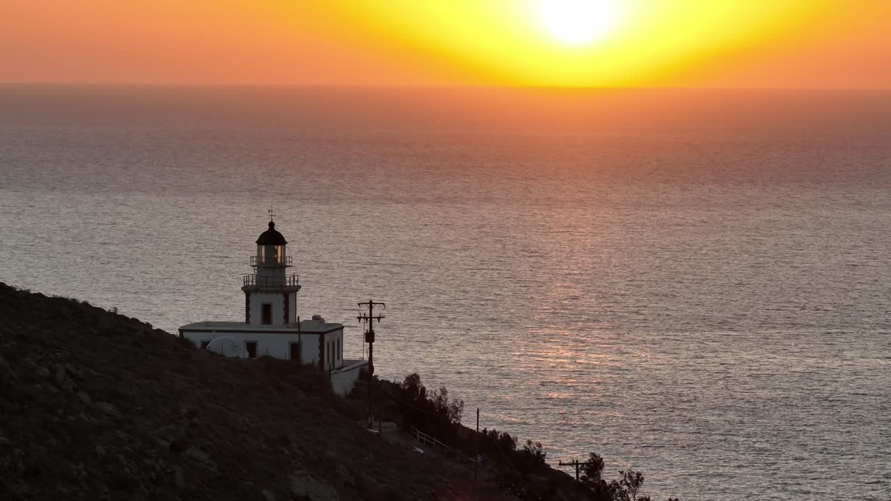 Drone revelando o farol de Santorini durante a hora dourada ao pôr do sol, capturando o horizonte, as falésias da caldeira e o profundo Mar Egeu azul em luz quente