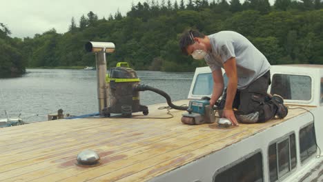 young man belt sanding planks on wooden boat forecabin