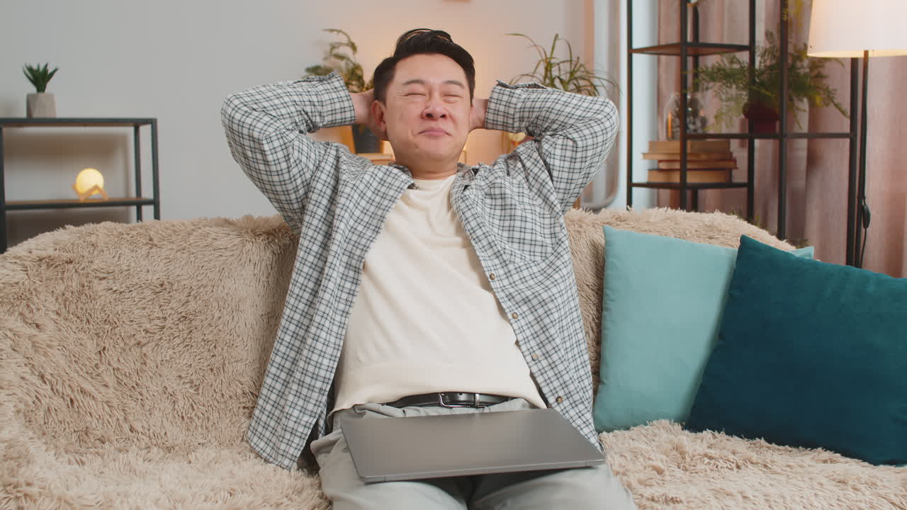 Happy mature chinese man sitting on couch closing laptop after finishing work in living room at home