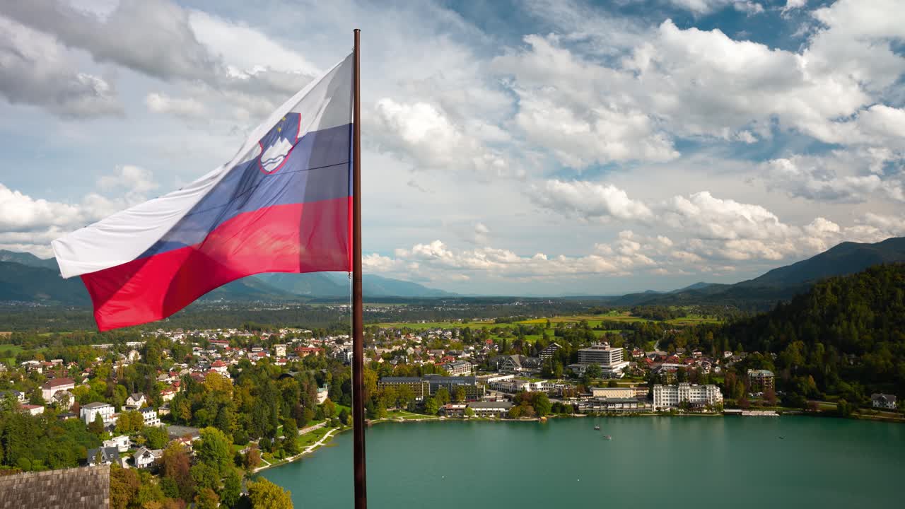 Slovinská vlajka vlající ve větru u hradu Bled. Zamračený podzimní den s horským pásmem v pozadí. Letecký detailní záběr. Modrá voda jezera a domy