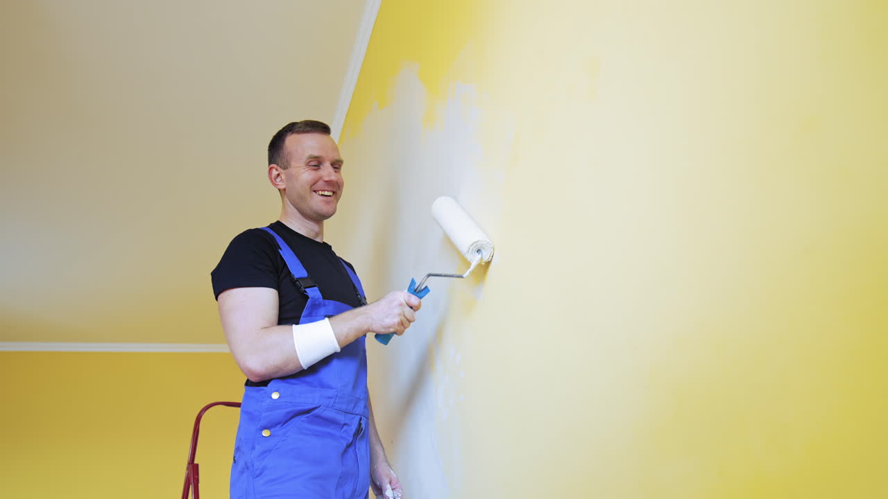 Painter with a paint roller in action. Happy young adult man painting the wall of the room in a flat. Interior decoration.