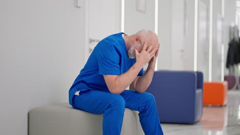 a sad and upset male doctor in a blue uniform an elderly man with glasses and a beard holds his head and cries in the corridor of a modern clinic. an experienced male doctor is upset and worried about his failure in his doctoral career