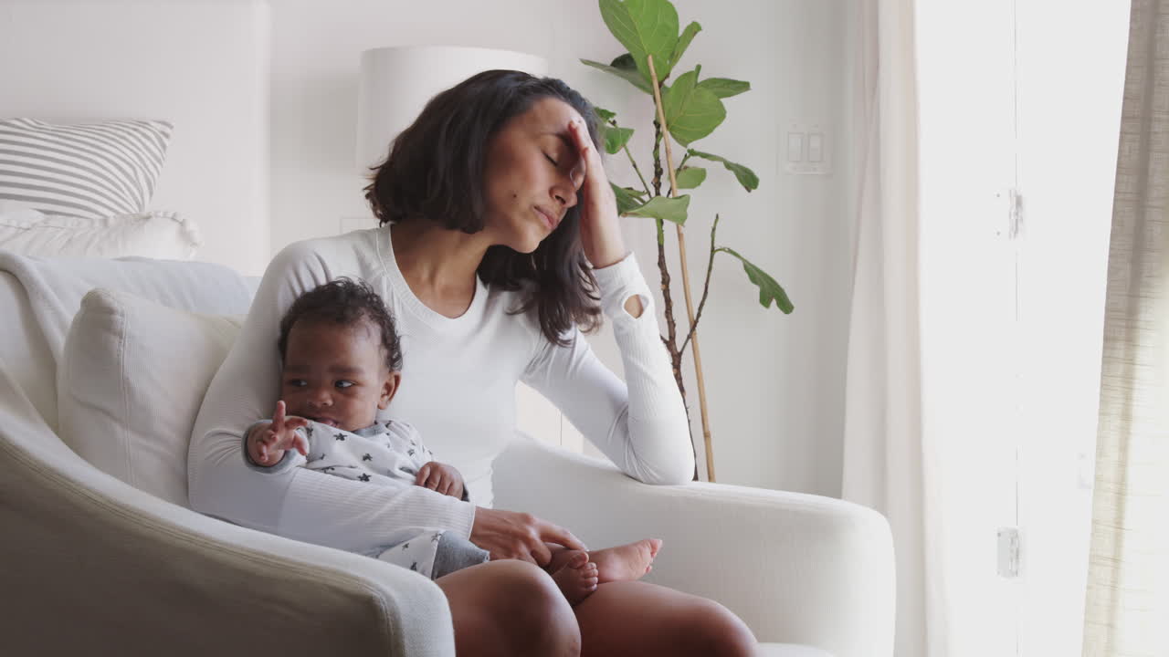 Tired young adult mother sitting in an armchair holding her baby and contemplating