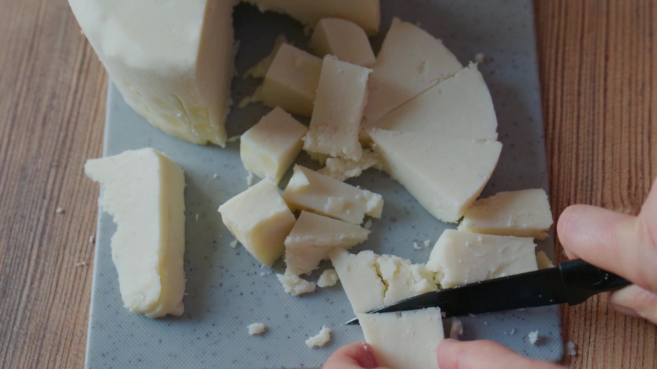 Primo piano di mani che affettano formaggio bianco a pasta dura su pietra grigia. Il coltello taglia una consistenza densa e cremosa. Pezzi di formaggio si spargono sul tagliere. Ideale per blog di cucina, video di ricette.