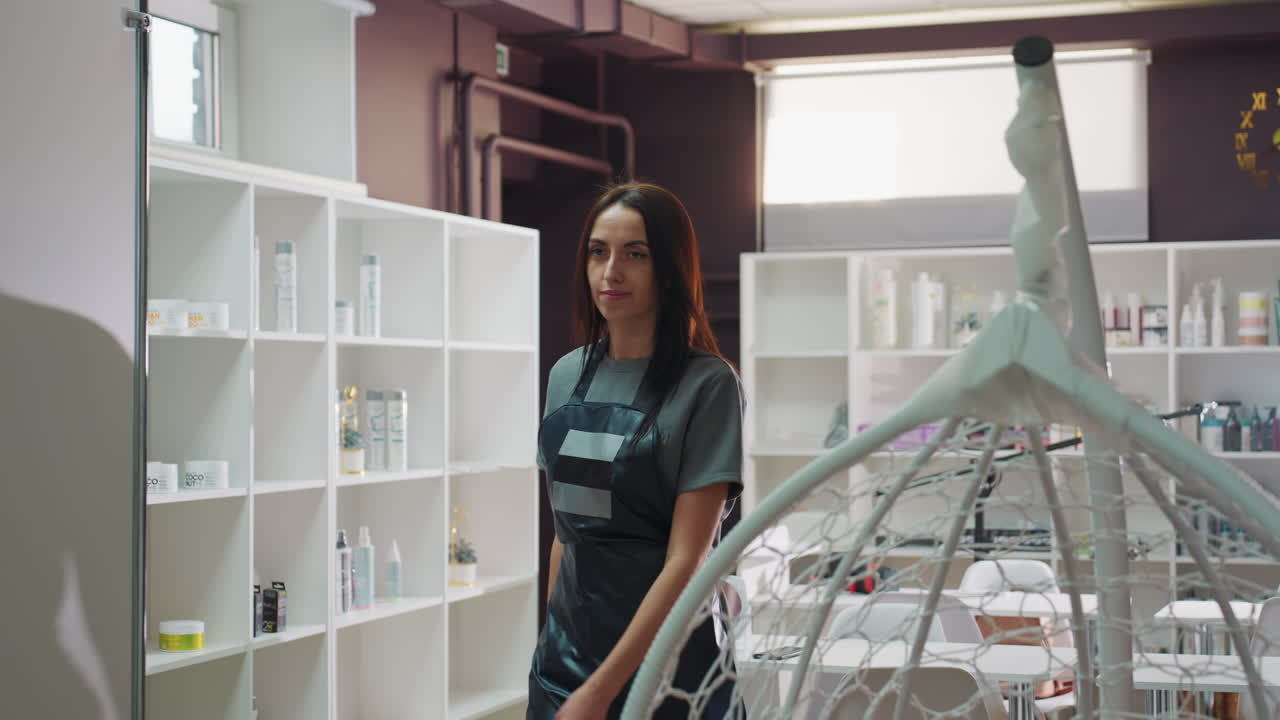 Una stilista caucasica single in piedi all'interno di una sala trattamenti sorride e saluta un cliente in arrivo; un grembiule professionale, scaffali di prodotti per la cura della pelle, una lampada clinica e attrezzature organizzate trasmettono l'idea di una spa moderna