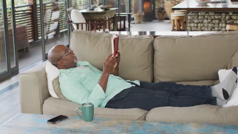 Glücklicher-Afroamerikanischer-älterer-Mann,-Der-Sich-Entspannt,-Auf-Der-Couch-Liegt-Und-Ein-Buch-Liest