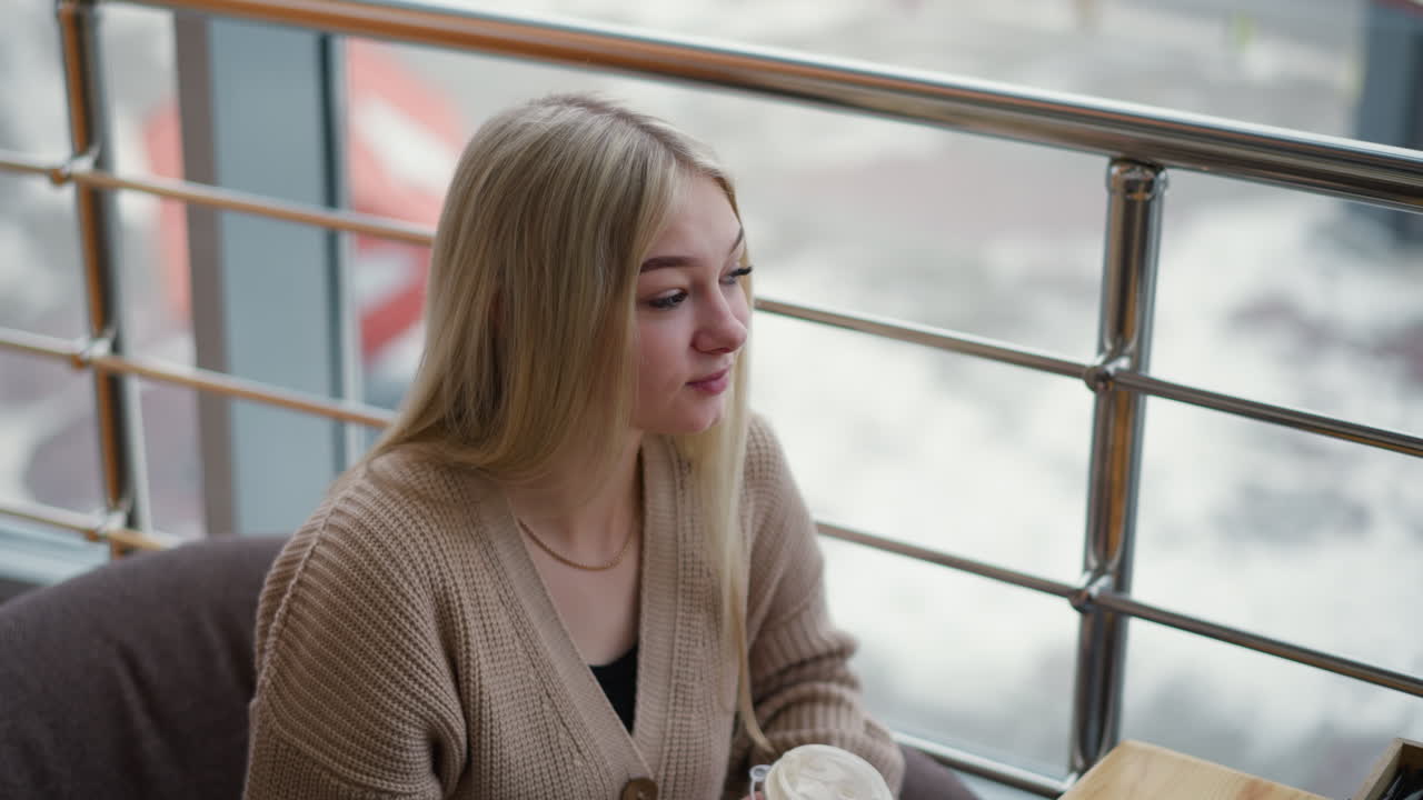 Signora pensierosa che sorseggia caffè mentre si guarda intorno con espressione rilassata, sullo sfondo di un paesaggio urbano invernale sfocato visibile attraverso la finestra, atmosfera accogliente in un moderno ambiente da caffè.