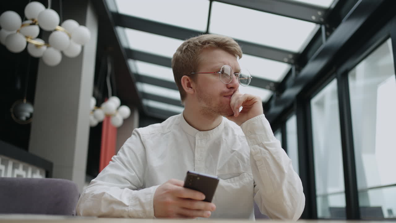A young man with glasses with a mobile phone in his hands looks at the screen and prints a message. Hipster in round glasses writes a message on his phone