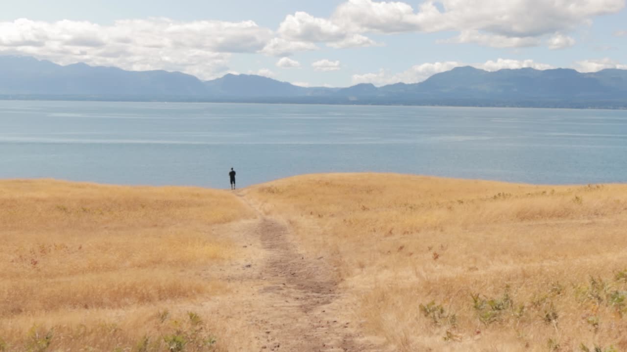 캐나다 브리티시 컬럼비아의 혼비 섬에 있는 아름다운 황금 초원과 산이 있는 바다가 내려다보이는 헬리웰 초원 난간 가장자리에 서 있는 남자