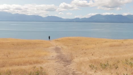 Man-standing-on-the-edge-of-the-Helliwell-meadow-ledge-overlooking-the-ocean-with-a-beautiful-golden-meadow-and-mountains-on-Hornby-Island-in-British-Columbia,-Canada