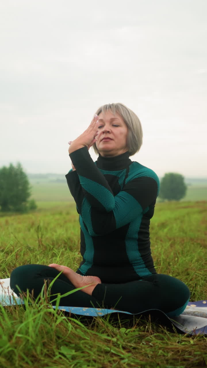 Woman seated on yoga mat in misty grassy field, twisting her hands while practicing yoga with eyes closed, under a cloudy sky, surrounded by nature in a vast serene landscape