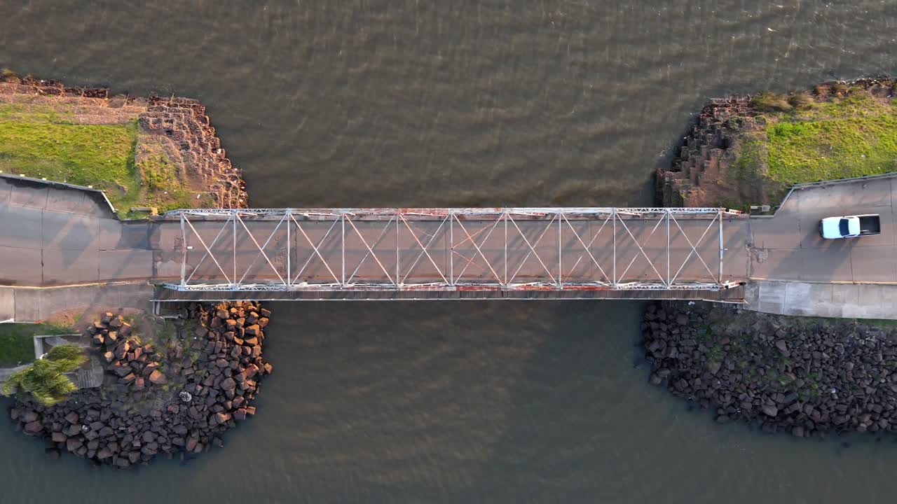 Aerial cinematic shot of a bridge over water with a red car and white van crossing; shadows cast by the evening light create a striking contrast.