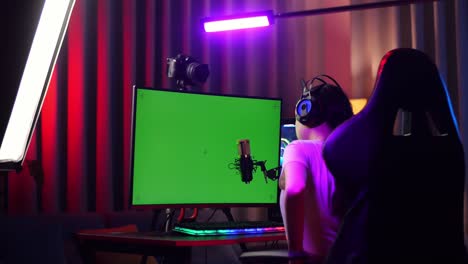 back view of asian girl streamer having a backache while using green screen personal computer. live stream on desk illuminated by rgb led strip light