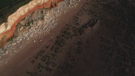 Aerial-Drone-Shot-Flying-Backwards-from-The-Wreck-Of-The-Steam-Trawler-Sheraton-Old-Shipwreck-on-Old-Hunstanton-Beach-with-Orange-and-White-Layered-Cliffs-at-Sunset-in-North-Norfolk-UK