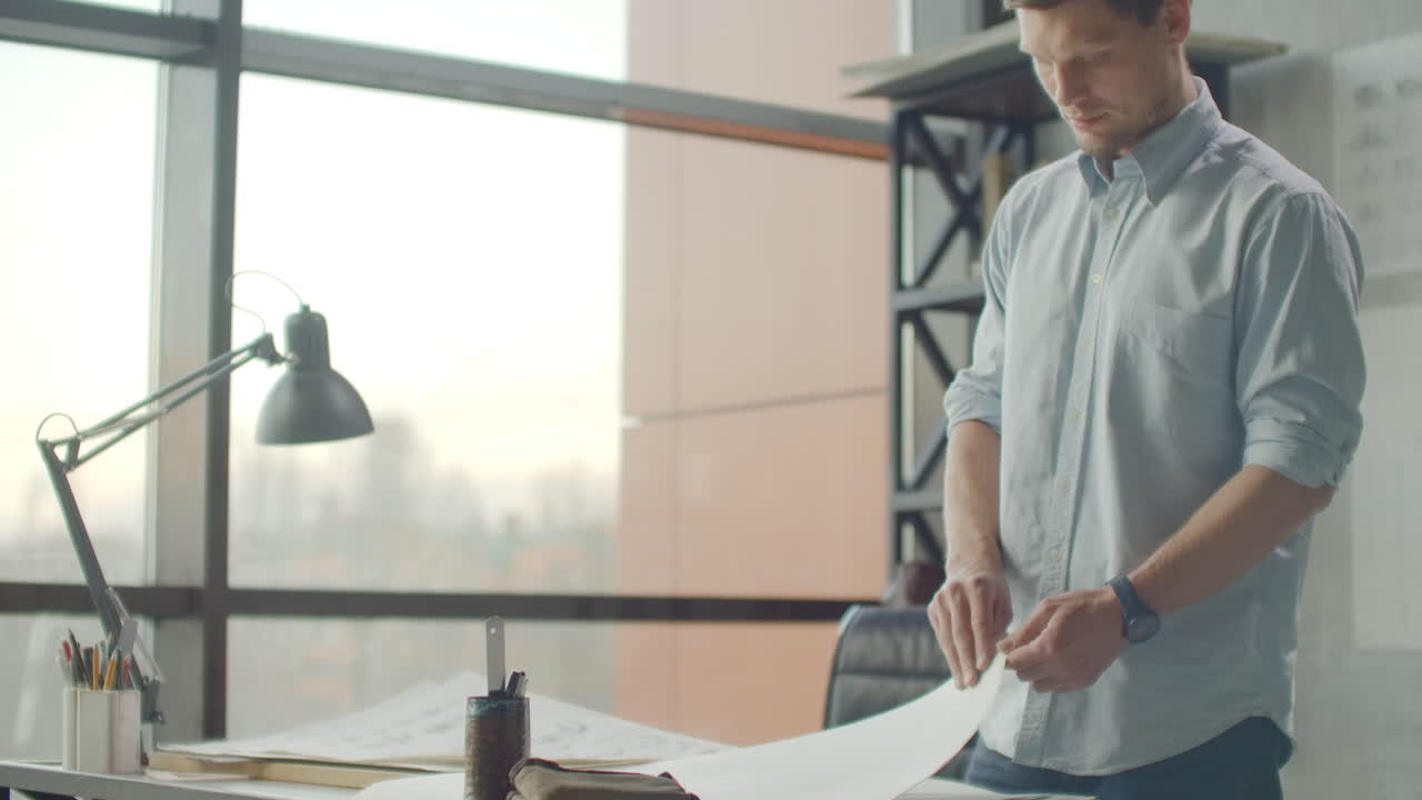 Hipster engineer works with blueprint. Shoot from above. Young architectural engineer working in office. Bearded man makes sketches. Loft style minimalistic interior drawings on the table