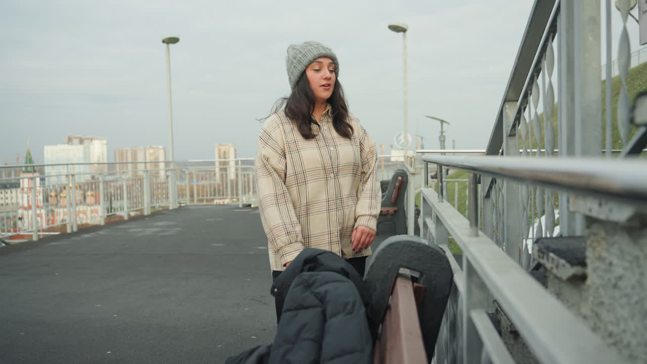 Donna bianca balla giocosamente su un ponte, con maglione spesso e berretto di lana. Sfondo dello skyline e luce coperta. Un pendolare attraversa con passo deciso. Un artista improvvisato che si lancia in...