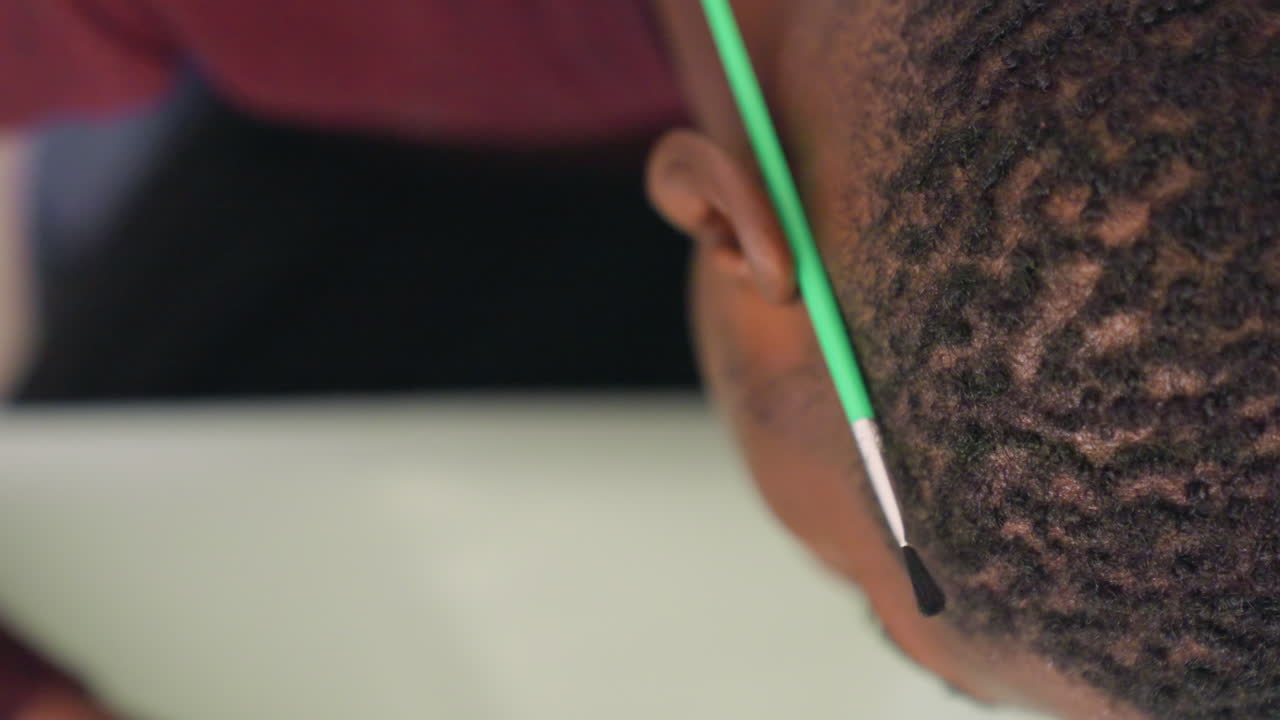 Pittura a pennello di capelli neri in primo piano, scena di classe tranquilla con giovane studente seduto al tavolo, la punta delicata del pennello che traccia il cuoio capelluto e l'attaccatura dei capelli, onde e riccioli texturizzati catturati in luce soffusa, attento.
