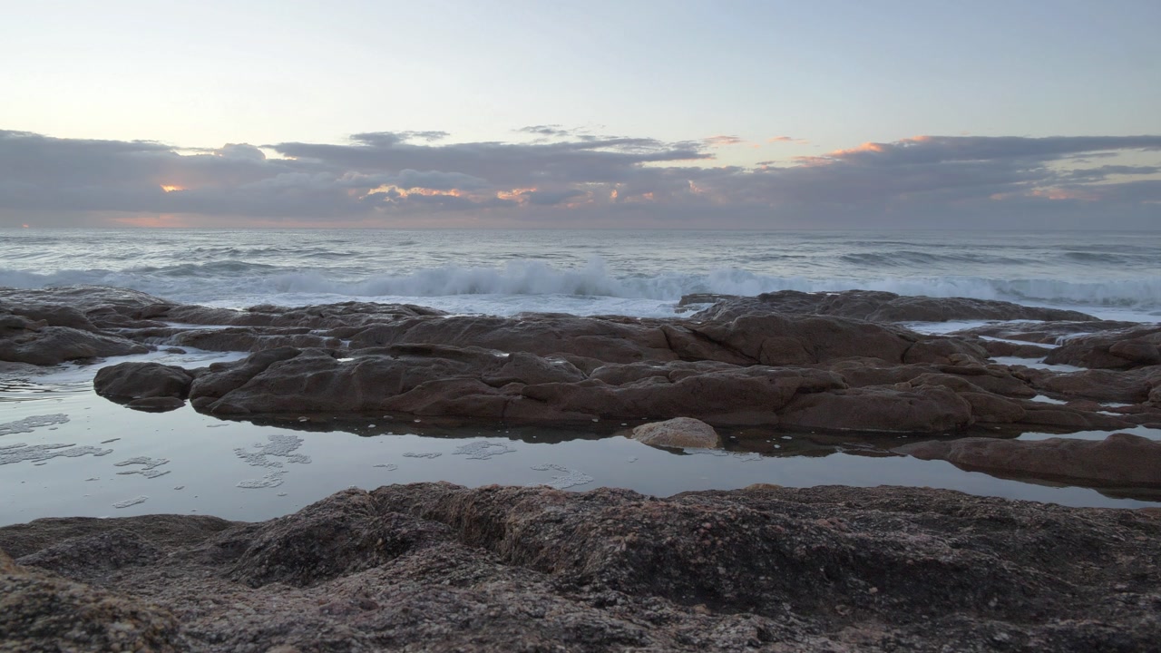 Gentle ocean waves roll onto a rugged coastline with tide pools reflecting the soft hues of the early morning sky at Ramsgate, South Coast.