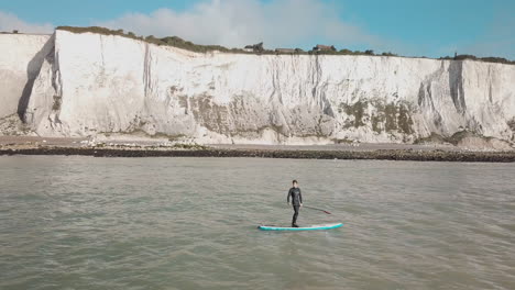 un joven se pone de pie haciendo paddle surf en el mar con los acantilados blancos de dover al fondo