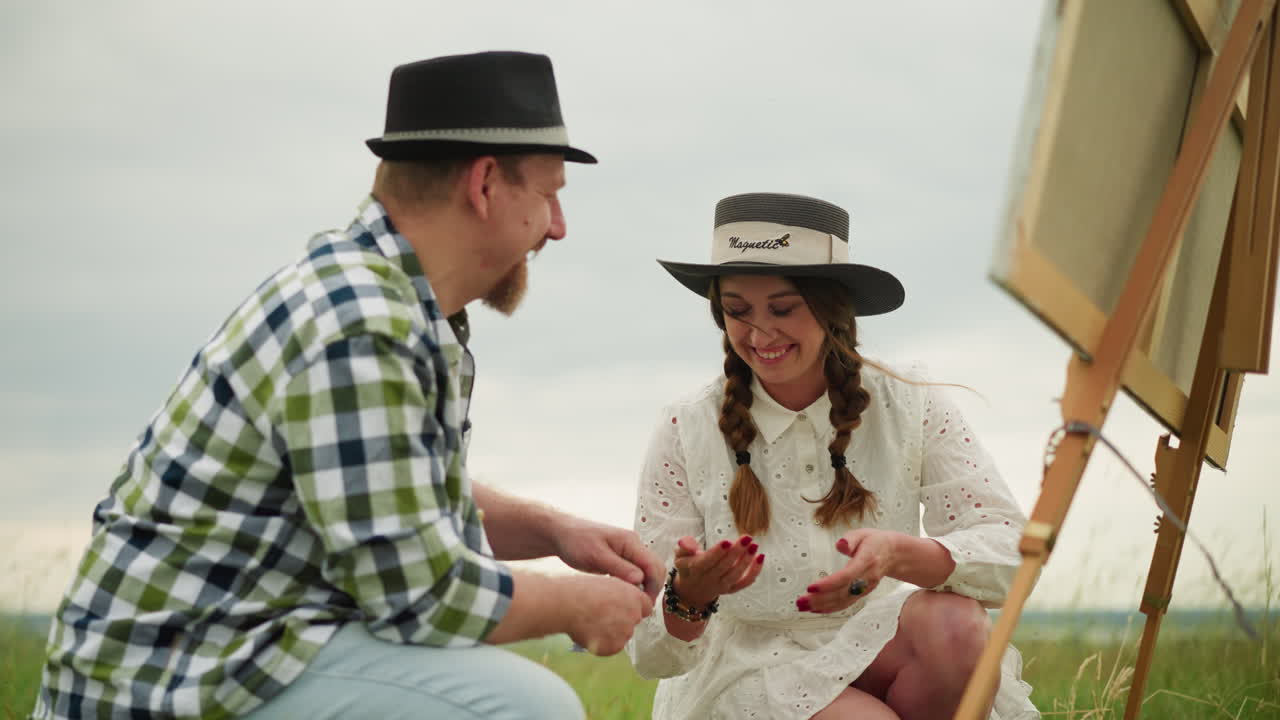 색 셔츠와 모자를 입은 장인 한 명이 페인트 튜브를 열기 위해 고군분투하는 동안, 색 드레스와 모자 입은 여자가 그의 노력에 웃는 유머러스한 장면