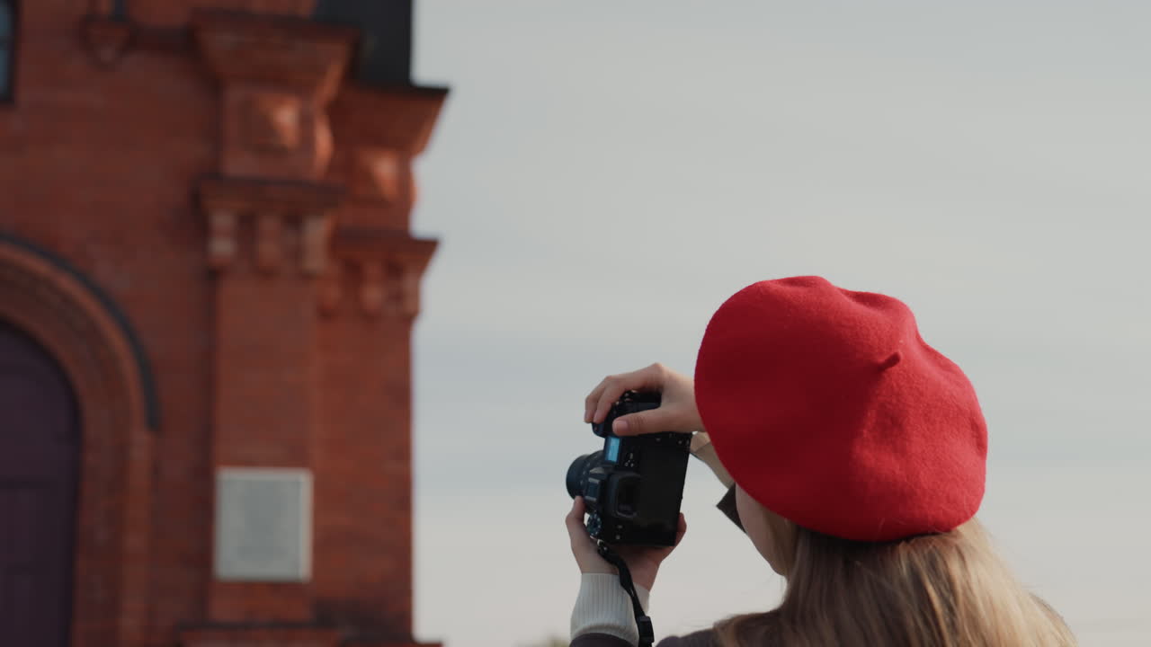 Donna bianca con berretto rosso che fotografa una chiesa, catturando la facciata in mattoni rossi e la cupola dorata. Fotografa di strada e studentessa di architettura che inquadra dettagli di campanile e archi, con in mano una macchina fotografica vintage, cappotto