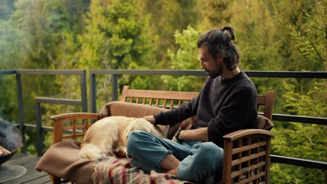 a young brunette man pets a large light-colored dog on a sofa on the balcony of a country house overlooking the mountains and pine forest