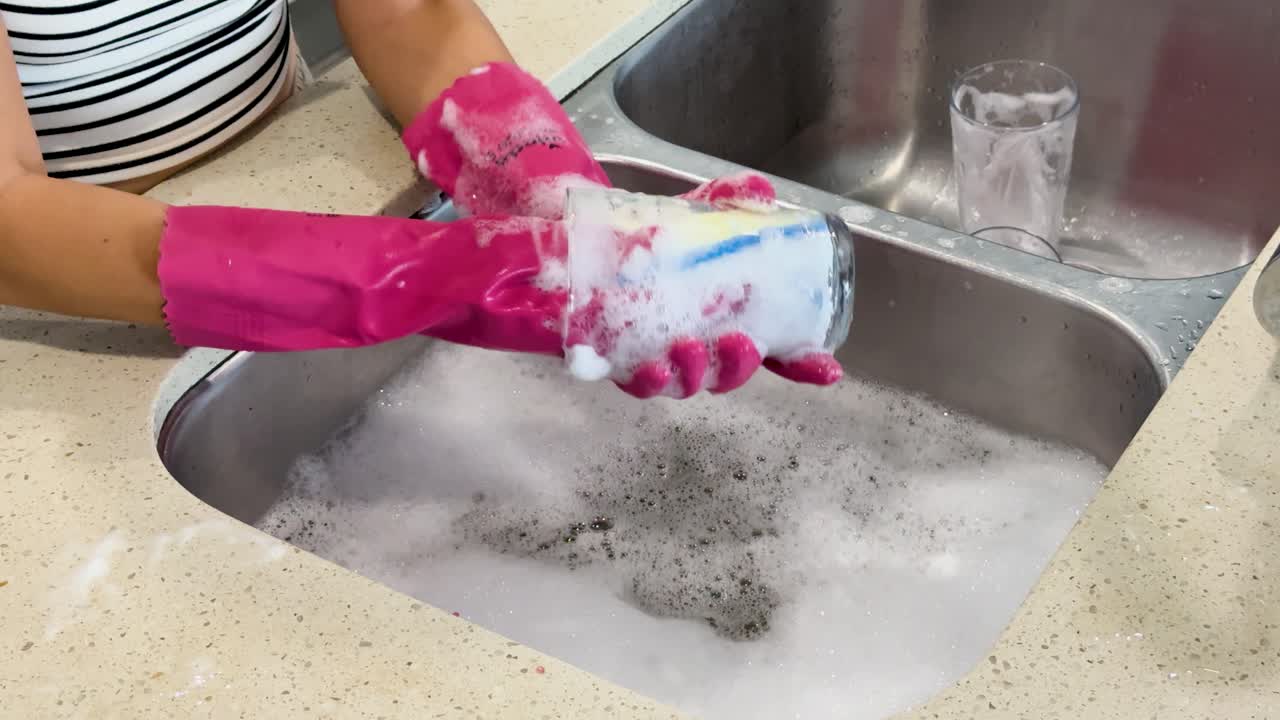 Individual wearing pink rubber gloves thoroughly scrubs a glass cup with a sponge in a modern kitchen sink, using soapy water and bright lighting