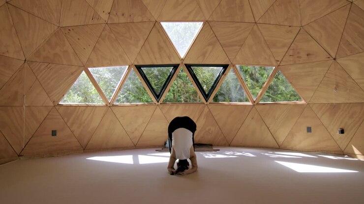 Young woman performing yoga stretches in a peaceful geometric outdoor studio, stretching forward into headstand at center of room
