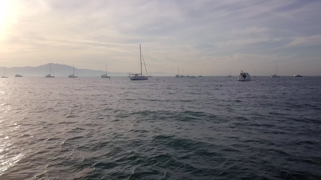 A wide coastal view showing boats spread across gentle ocean waves under a pale morning sky, with distant mountains forming the horizon