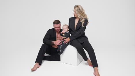 beautiful blonde woman sits on white cube holding her baby. man sits beside the cube playing with child. studio footage. white backdrop.