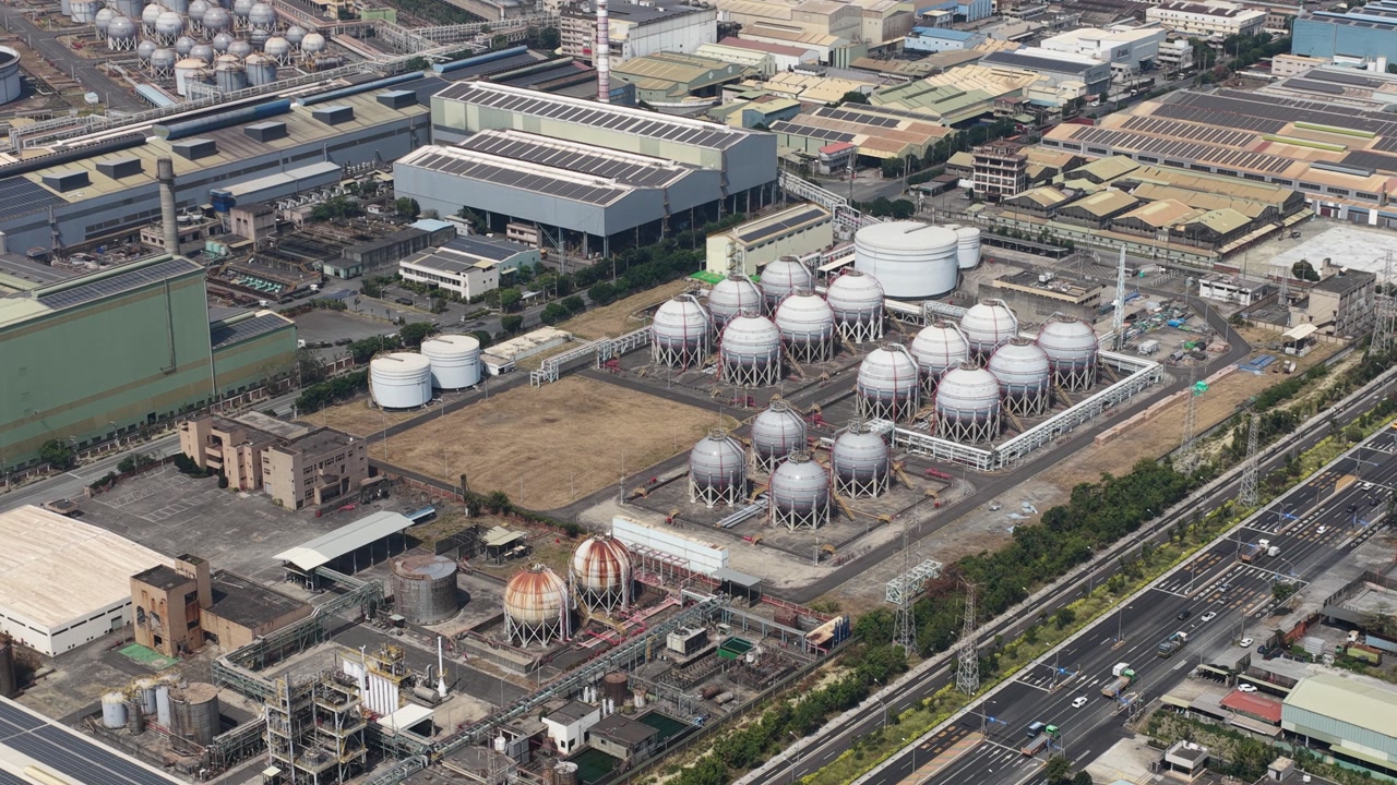 Expansive aerial perspective showing a large industrial complex with numerous spherical gas storage tanks and a busy highway running alongside, highlighting energy production and infrastructure