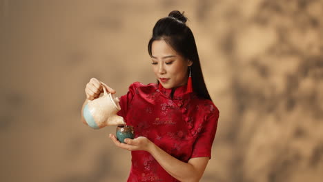 young asian woman in red traditional clothes pouring hot tea from ceramic kettle to cup and smiling at camera