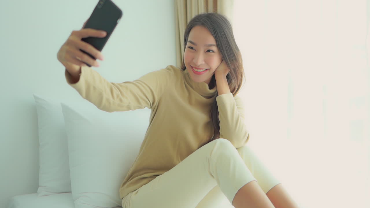 Smiling Happy Young Woman sitting on bed taking selfie photos, Morning lights
