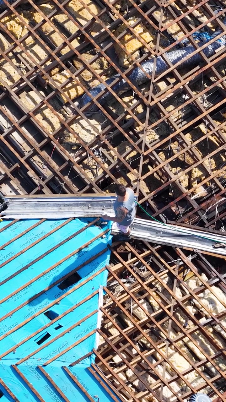 Top-down drone shot of workers installing blue sarking over timber trusses during house renovation.