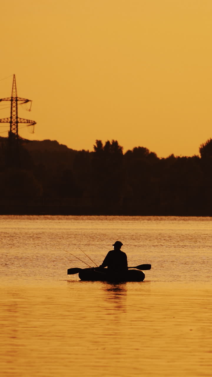 Silhouette of a man floating in a boat on the river surface on the background of an evening trees. Fisherman roams with oars while sailing on a row boat in the lake at beautiful sunset. Vertical video