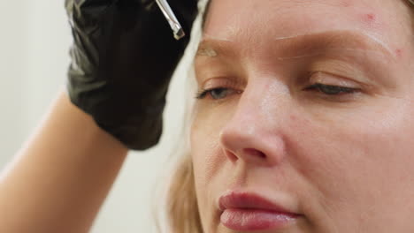 close-up view of technician gently applying cream to client eyebrow using precision brush during beauty session, with client eyes partially closed and gloved hand holding tool near eyebrow outline