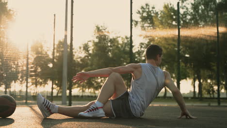 Konzentrierter-Männlicher-Basketballspieler,-Der-Sich-Dehnt-Und-Sitzt,-Sitzt-Auf-Dem-Boden-Im-Basketballplatz-Im-Freien